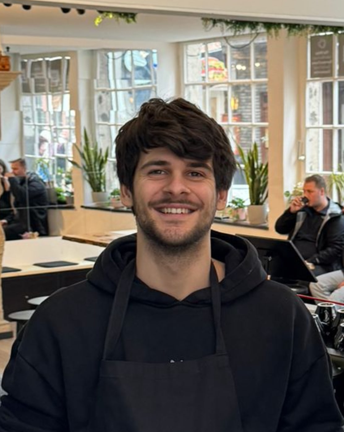 Barista Merlin lächelt in die Kamera, im Hintergrund ein Café mit anderen Gästen.