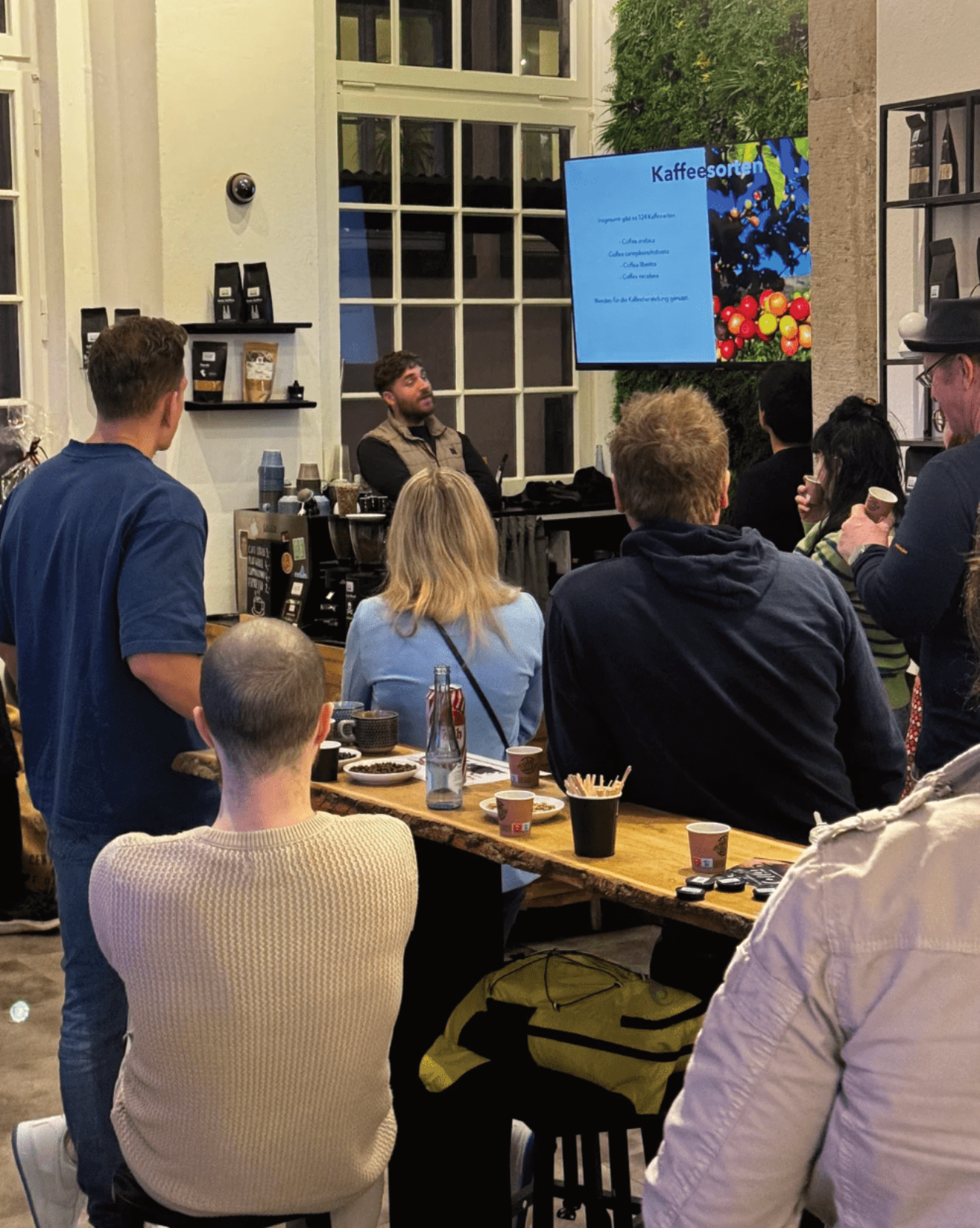 Eine Gruppe von Menschen hört einem Vortrag über Kaffeesorten in einem Café zu. Ein Redner steht vor einem Bildschirm, auf dem Informationen über verschiedene Kaffeesorten angezeigt werden.