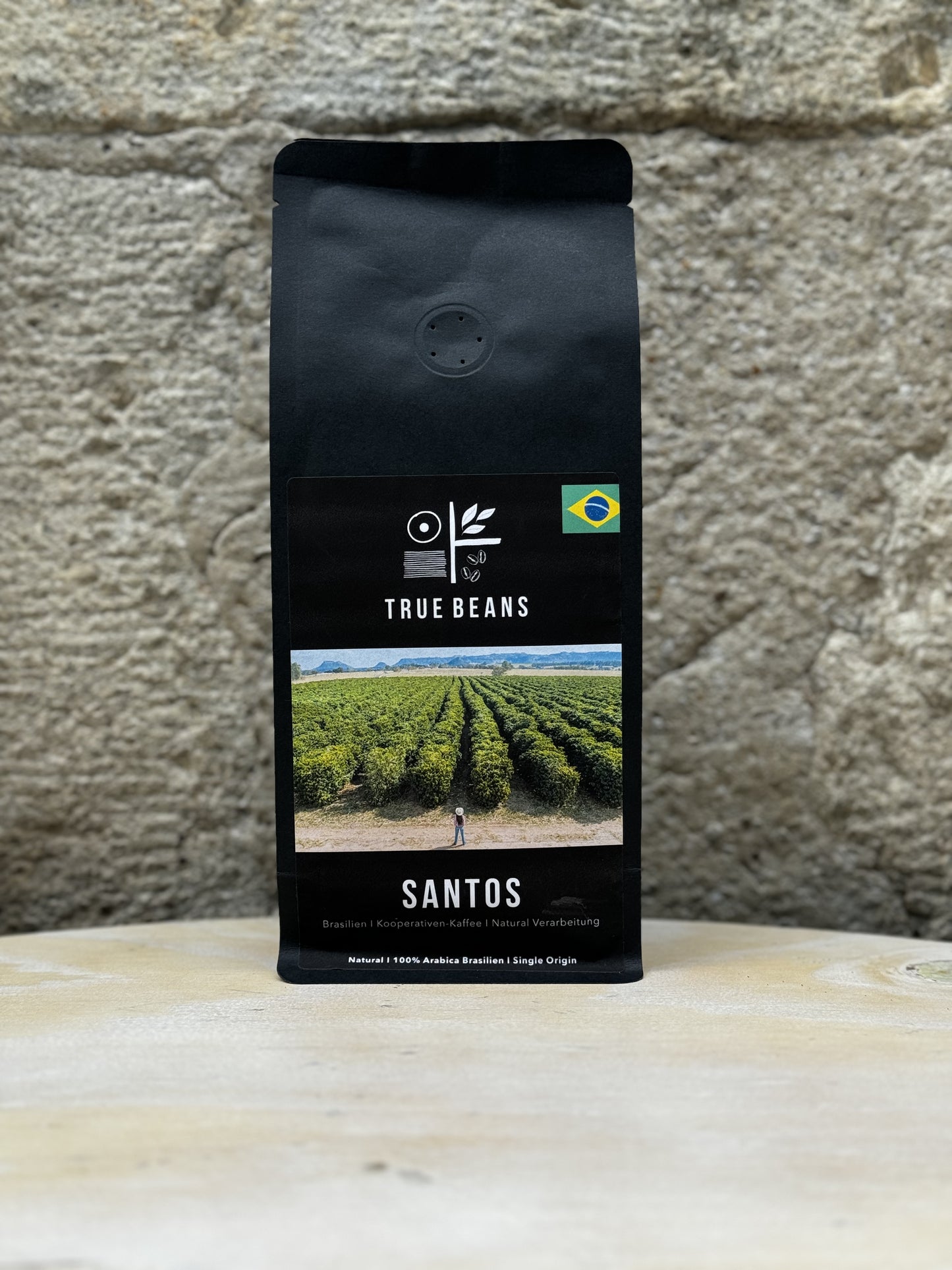 Eine Packung True Beans Santos Kaffee vor einer Steinwand, mit der Aufschrift "Brasilien | Kooperativen-Kaffee | Natural Verarbeitung" und einem Bild einer Kaffeeplantage mit einem Arbeiter inmitten der Felder.