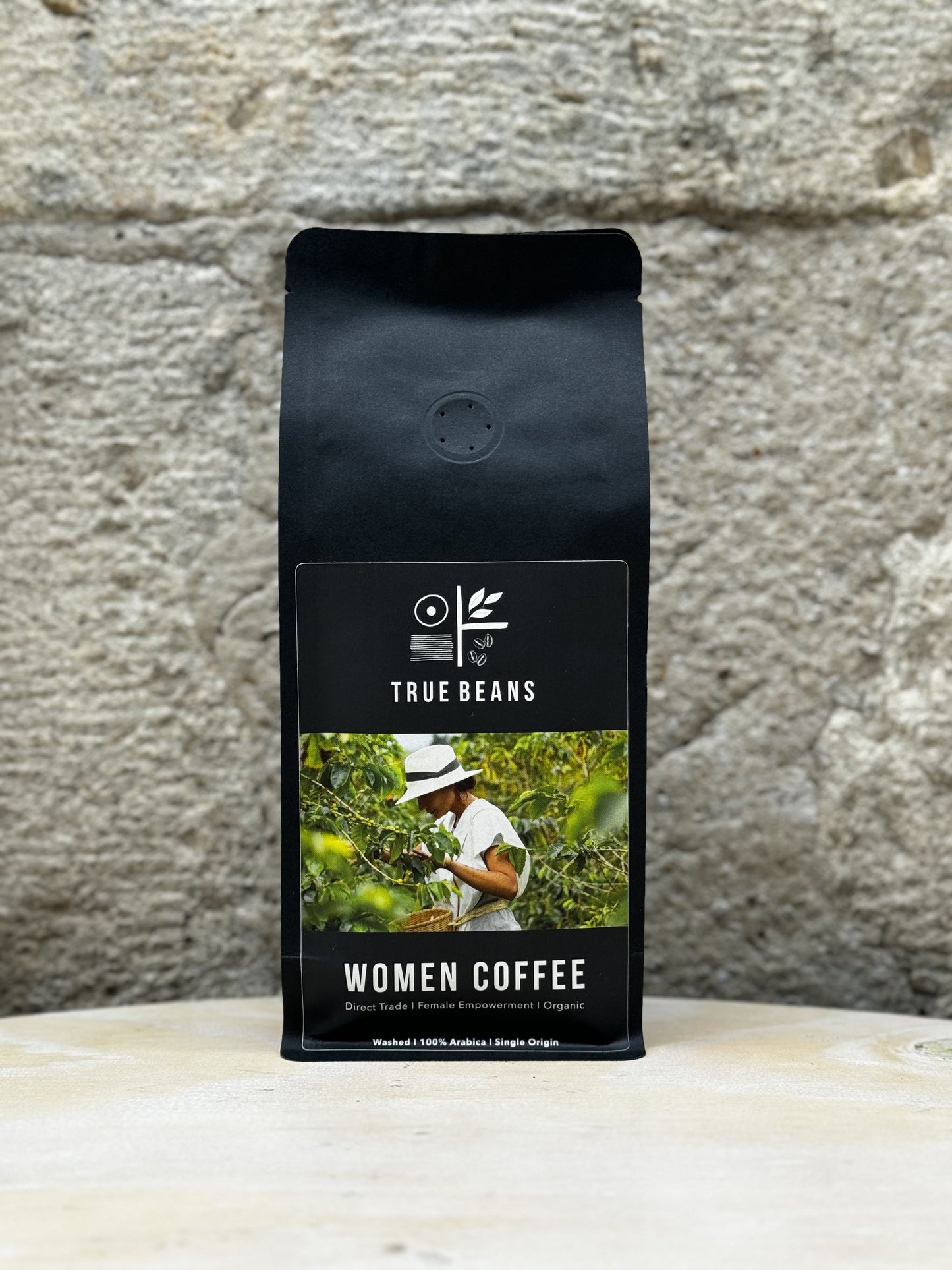 Eine Packung True Beans Women Coffee vor einer Steinwand. Auf der Verpackung ist ein Bild einer Frau, die Kaffee pflückt, sowie die Beschreibung "Direct Trade | Female Empowerment | Organic" zu sehen.