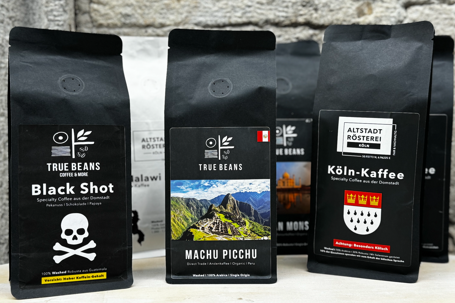 Eine Auswahl von Kaffeeverpackungen von True Beans und Altstadt Rösterei. Von links nach rechts sind die Sorten "Black Shot", "Machu Picchu" und "Köln-Kaffee" zu sehen. Jede Verpackung hat ein individuelles Design und Logo.