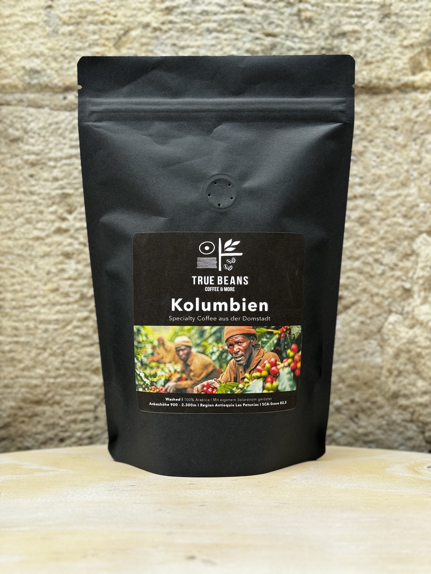Eine Packung True Beans Kaffee "Kolumbien" vor einer Steinwand, mit der Aufschrift "Specialty Coffee aus der Domstadt". Auf dem Etikett sind Kaffeebauern bei der Ernte abgebildet. Der Kaffee ist 100% Arabica und wurde in der Region Antioquia Las Petunias angebaut.
