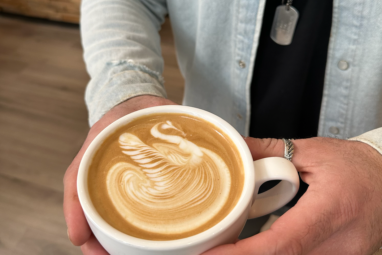 Eine Person hält eine Tasse Latte mit kunstvollem Schwan-Muster im Milchschaum, im Hintergrund ist ein Café zu sehen.