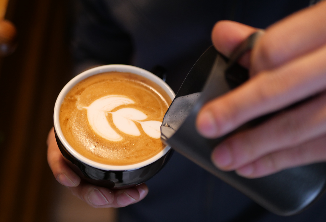 Ein Barista gießt Milch in eine Tasse Cappuccino und kreiert dabei ein Latte-Art-Muster in Form einer Tulpe. Die Tasse ist schwarz und wird von der linken Hand des Baristas gehalten, während die rechte Hand die Milchkännchen schwenkt. Die Aufnahme ist nah und fokussiert auf die Latte-Art.