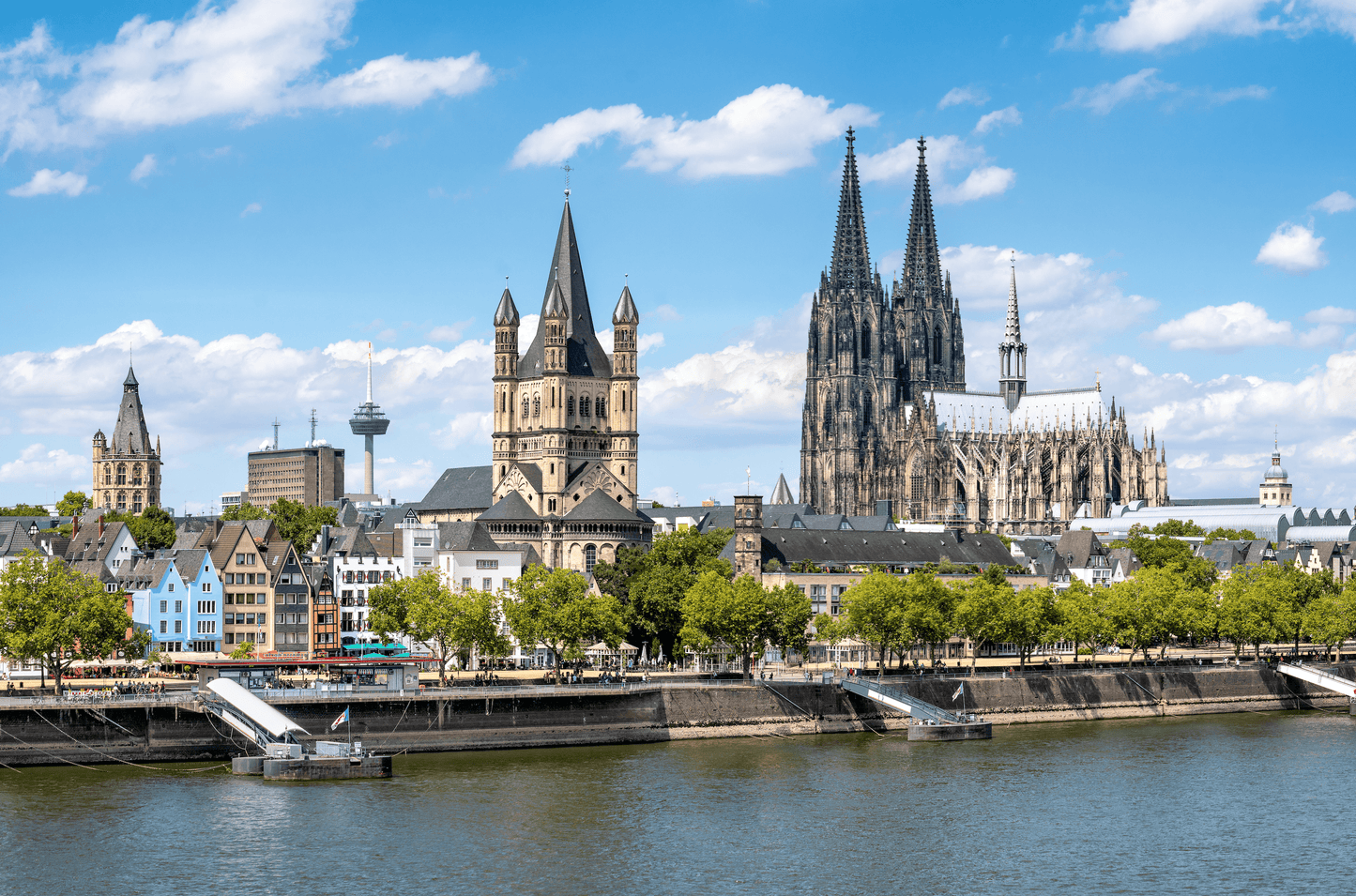 Panoramablick auf das Rheinufer in Köln mit dem Kölner Dom und der Kirche Groß St. Martin, im Hintergrund der Colonius Fernsehturm und andere städtische Gebäude unter einem blauen Himmel mit weißen Wolken.
