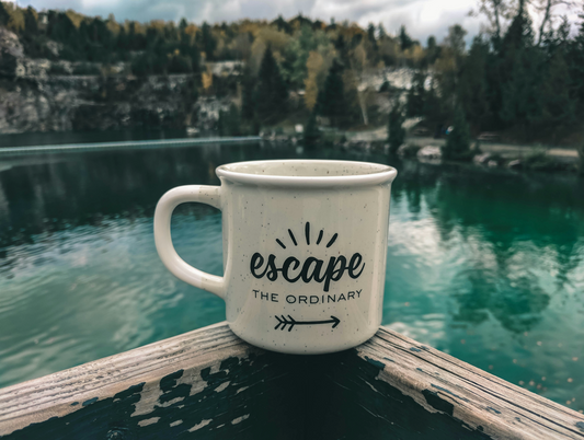 Tasse mit der Aufschrift "escape the ordinary" auf einem Holzbalken vor einem malerischen See und Wald im Hintergrund
