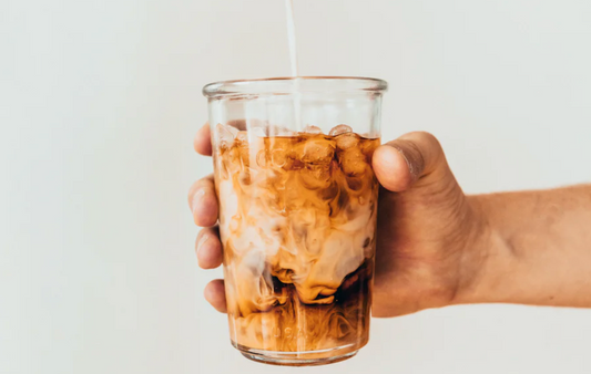 Ein Glas Eis Kaffee mit Milch, das in einer Hand gehalten wird, wobei die Milch sich malerisch mit dem dunklen Kaffee vermischt.