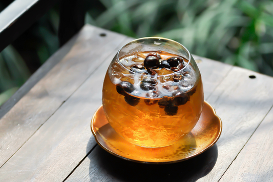 Ein gläserner Becher mit Kaffeekirschentee, gefüllt mit Eiswürfeln und schwimmenden Kaffeekirschen, steht auf einem hölzernen Tisch im Freien mit grünen Pflanzen im Hintergrund