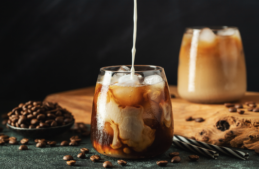 Kaffee Sirup wird in ein Glas mit Eiskaffee gegossen, umgeben von Kaffeebohnen auf einer dunklen Oberfläche.