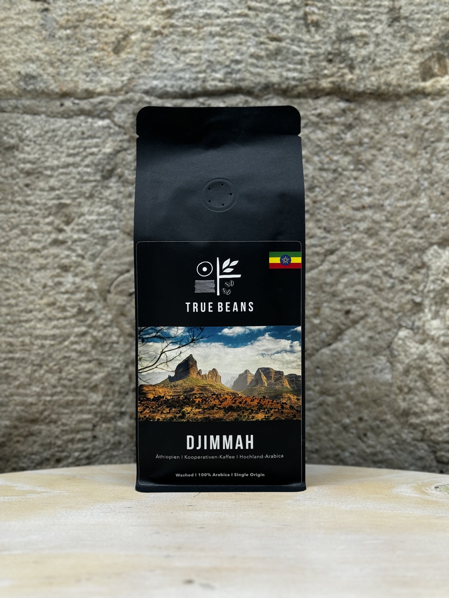 Eine Packung True Beans Kaffee "Djimmah" vor einer Steinwand, mit der Aufschrift "Äthiopien | Kooperativen-Kaffee | Hochland-Arabica". Auf dem Etikett ist eine malerische Landschaft mit Bergen und einem dramatischen Himmel abgebildet. Der Kaffee ist 100% Arabica und wurde gewaschen verarbeitet.