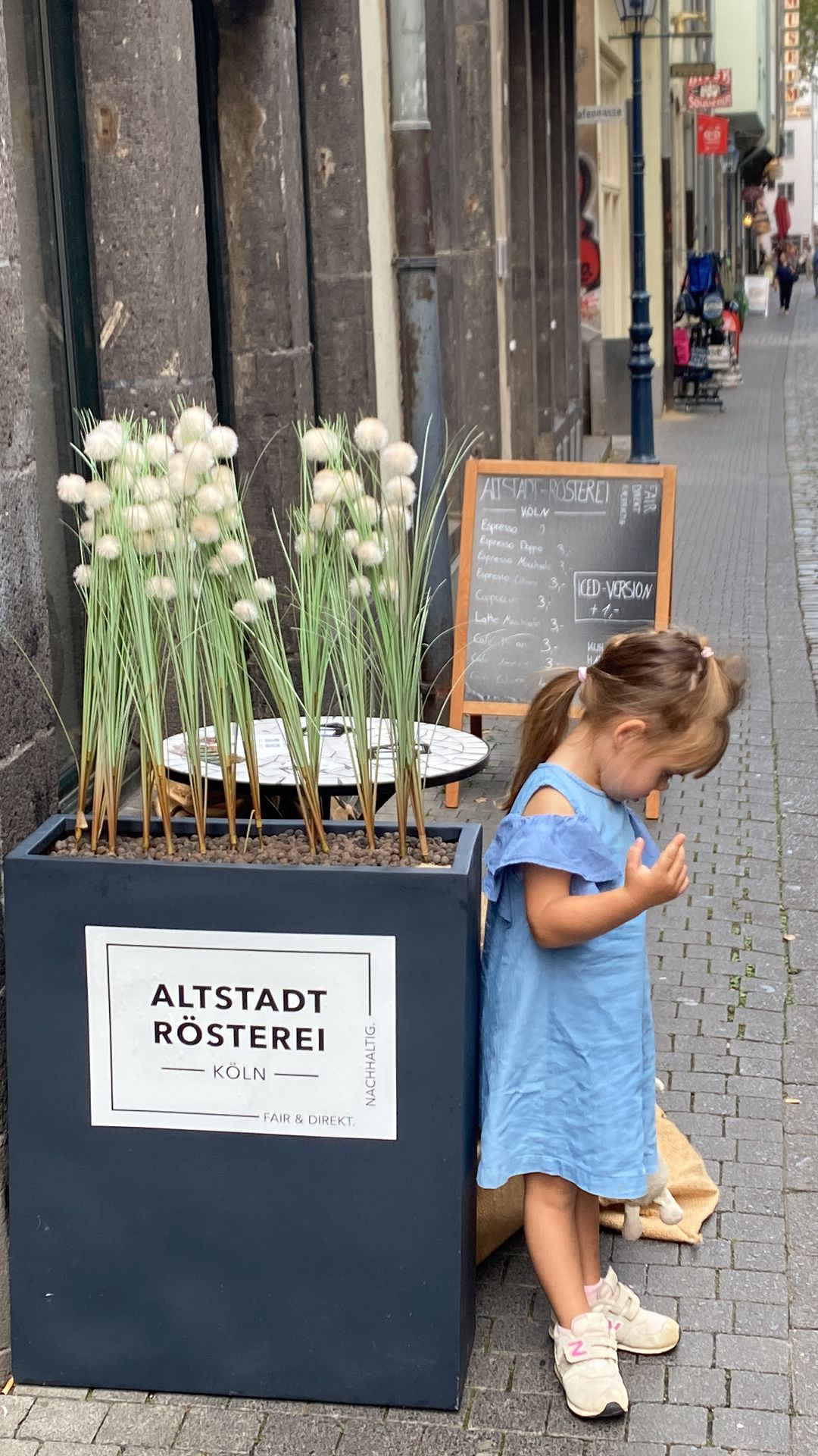 Ein kleines Mädchen in einem blauen Kleid steht neben einem großen Pflanzgefäß mit der Aufschrift "Altstadt Rösterei Köln" auf einer Straße in Köln. Im Hintergrund ist eine Kreidetafel mit dem Menü der Rösterei zu sehen.