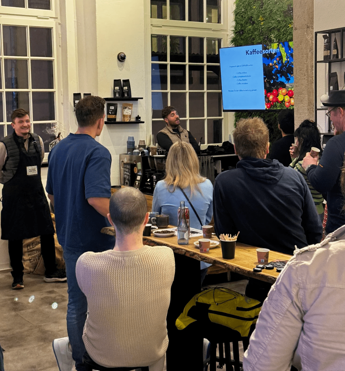 Eine Gruppe von Menschen hört einem Vortrag über Kaffeesorten in einem Café zu. Ein Redner steht vor einem Bildschirm, auf dem Informationen über verschiedene Kaffeesorten angezeigt werden.