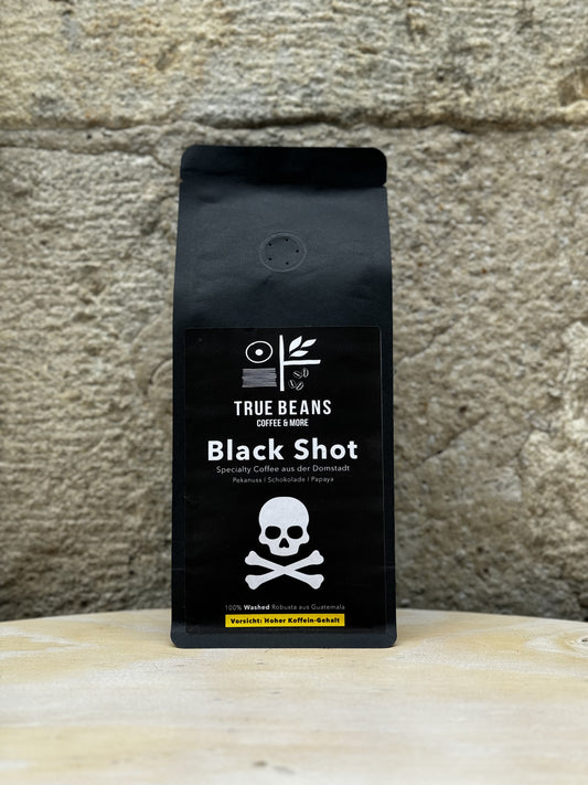 Eine Packung True Beans Black Shot Kaffee vor einer Steinwand. Auf der Verpackung ist ein Totenkopf mit gekreuzten Knochen abgebildet, sowie die Beschreibung "Specialty Coffee aus der Domstadt" und "100% Washed Robusta aus Guatemala". Darunter steht "Vorsicht: Hoher Koffein-Gehalt".