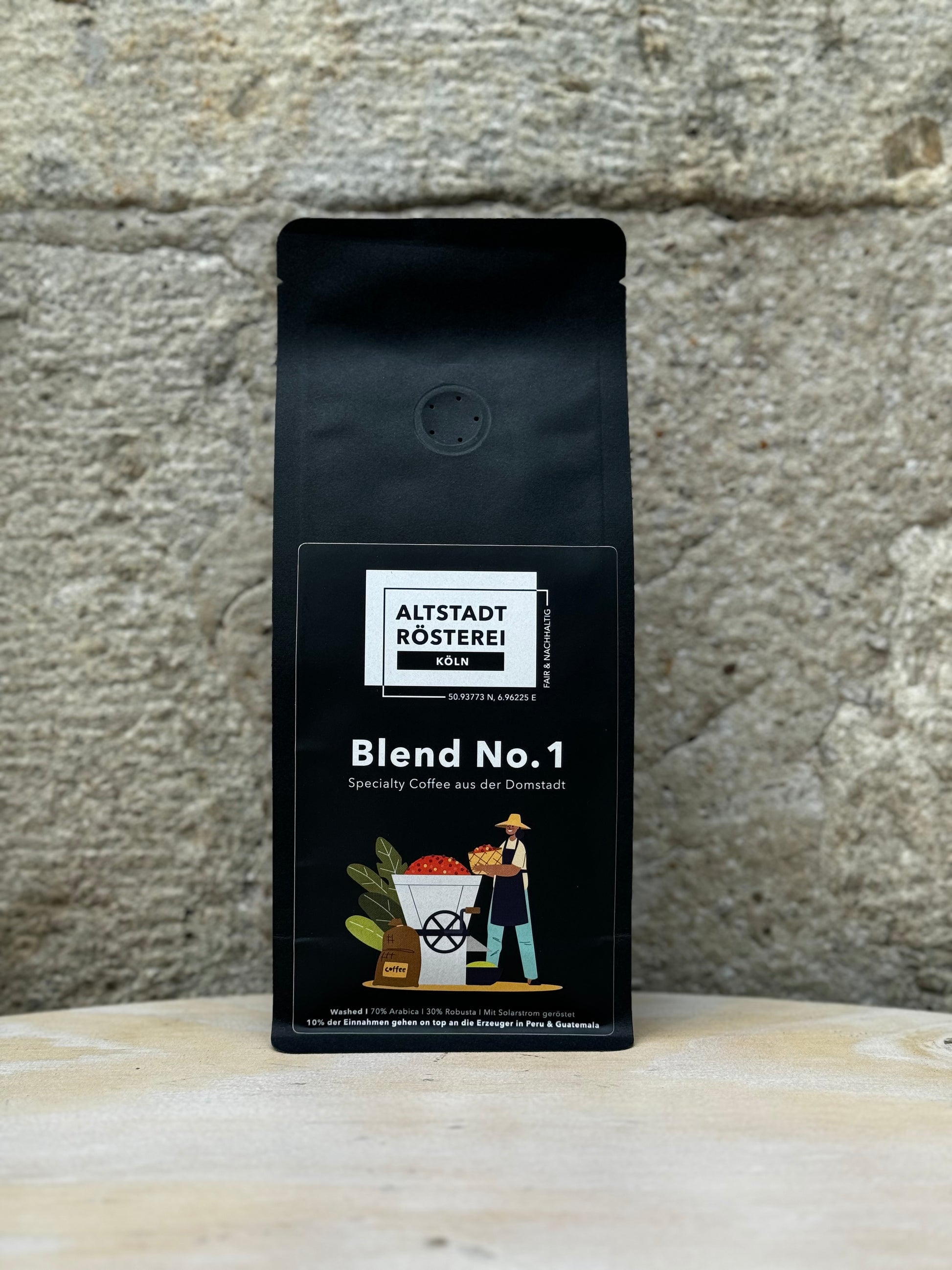 Eine Packung Altstadt Rösterei Köln Blend No. 1 Kaffee vor einer Steinwand. Auf der Verpackung ist eine Illustration eines Kaffeebauern mit Kaffeebohnen und Pflanzen zu sehen.