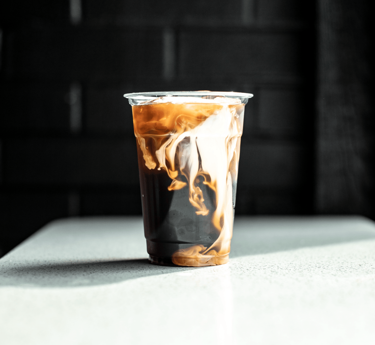 Ein Glas Cold Brew Kaffee, in dem sich langsam Milch oder Sahne einmischt, was zu einer faszinierenden, wirbelnden Optik führt. Die Kontraste zwischen dem dunklen Kaffee und der hellen Milch erzeugen ein ästhetisches und erfrischendes Bild. Der Hintergrund ist dunkel gehalten, was den Fokus auf das Getränk lenkt und die Details der Mischung hervorhebt.