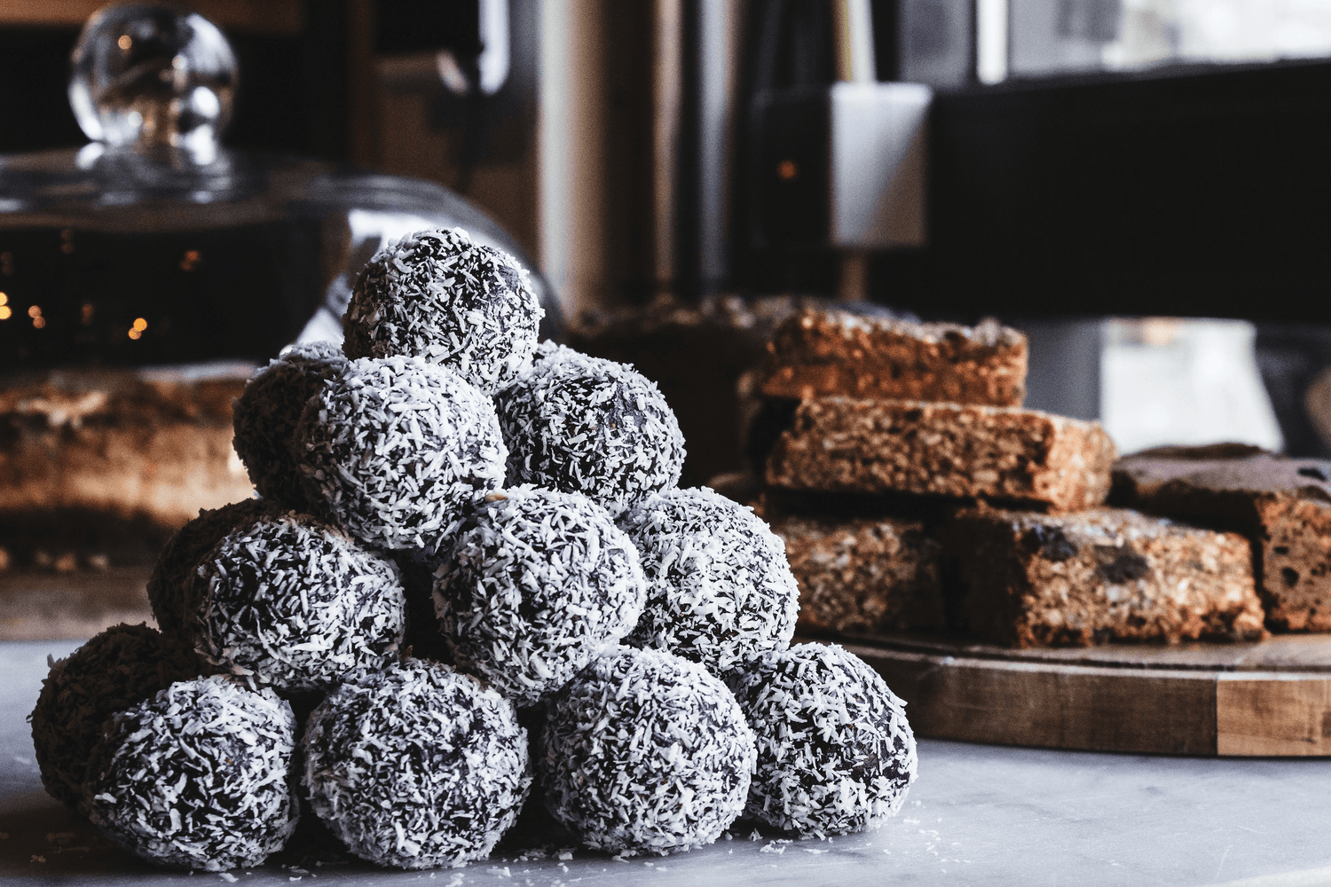 Ein Stapel von Kakao-Dattel-Bällchen, die mit Kokosraspeln bedeckt sind, steht im Vordergrund. Im Hintergrund sind verschiedene Gebäckstücke und Kuchen auf einem Tisch zu sehen. Die Szene vermittelt eine gemütliche Café-Atmosphäre.