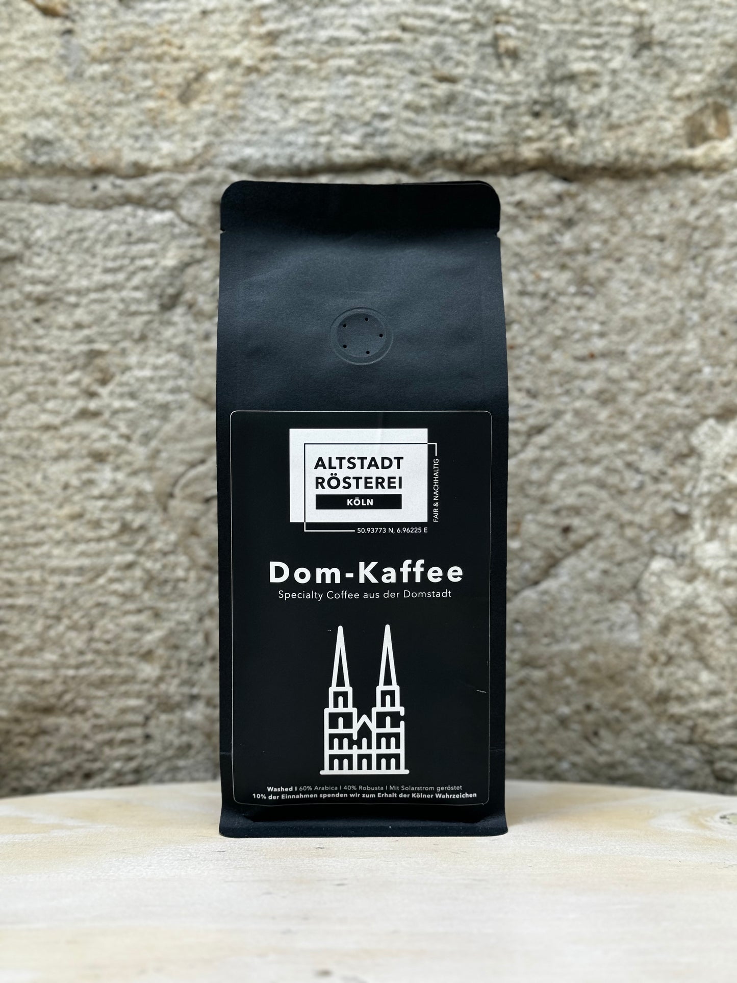 Eine Packung Altstadt Rösterei Dom-Kaffee vor einer Steinwand, mit der Aufschrift "Specialty Coffee aus der Domstadt" und einer Abbildung des Kölner Doms.