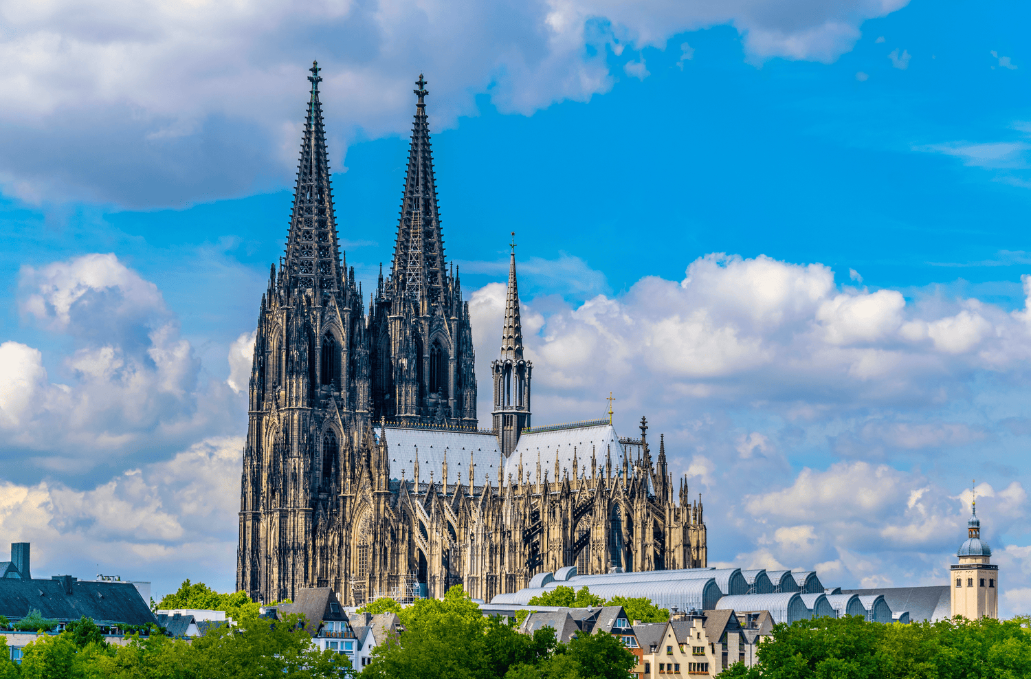 Das Bild zeigt den Kölner Dom bei Tageslicht mit kleinen Wolken am Himmel im Hintergrund. Die gotische Kathedrale ist gut beleuchtet und die Details der Fassade sind deutlich sichtbar.