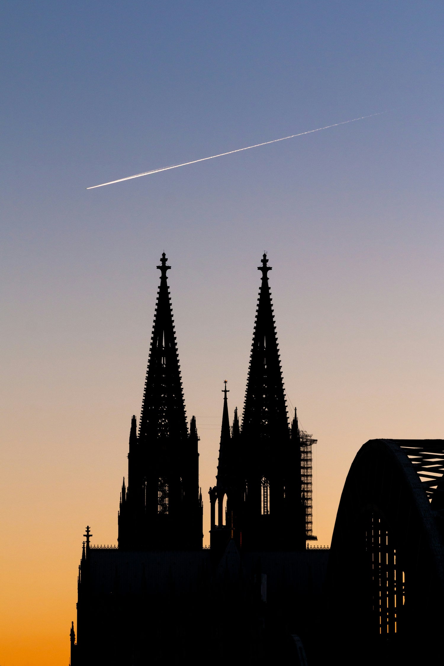 Silhouette des Kölner Doms bei Sonnenuntergang, mit einem Kondensstreifen eines Flugzeugs, das über den klaren Himmel fliegt.