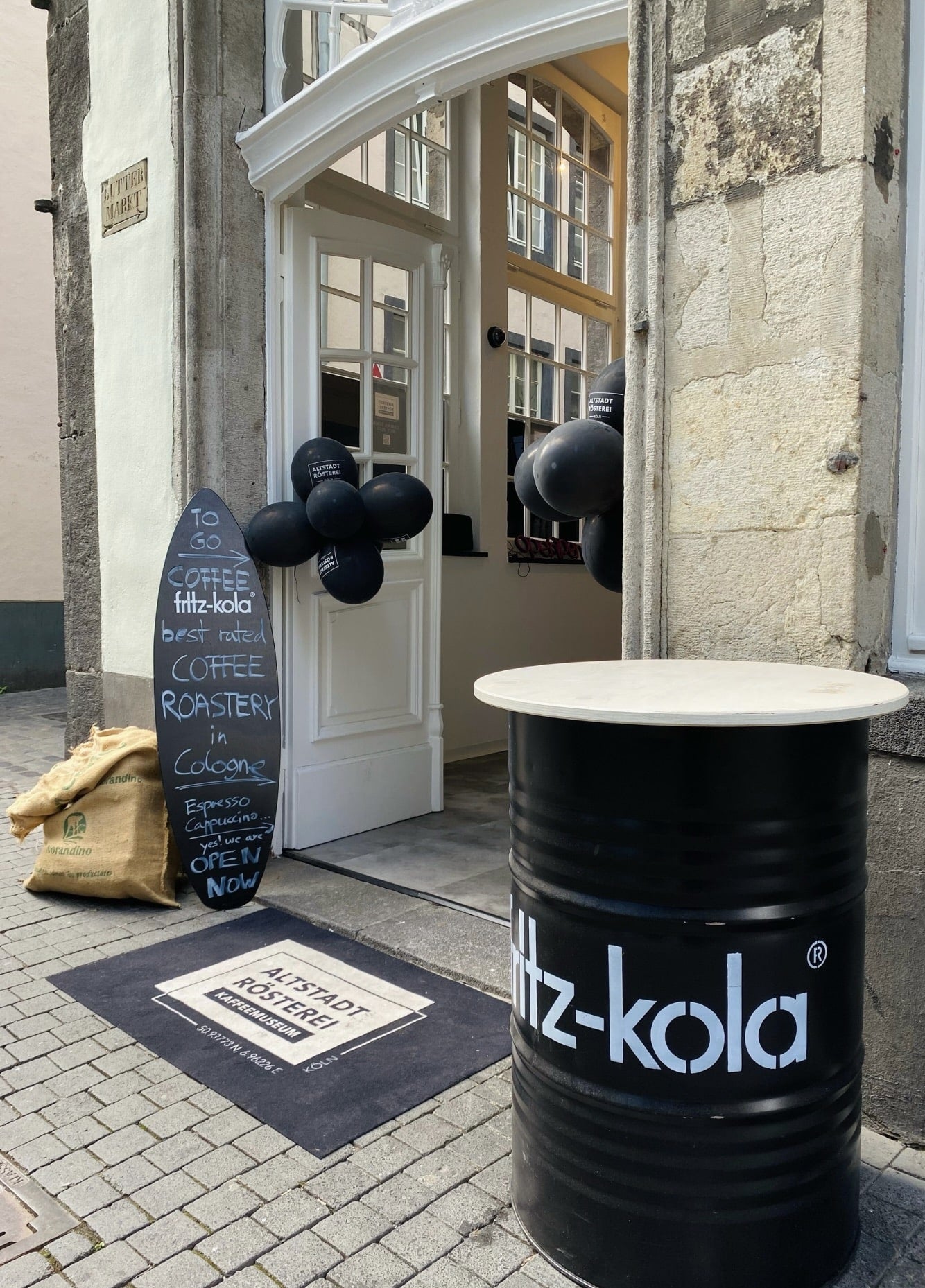 Der Eingang zur Altstadt Rösterei in Köln, dekoriert mit schwarzen Luftballons und einem Schild, das Kaffee und fritz-kola anbietet. Vor dem Eingang steht ein Tisch aus einem umfunktionierten Fass mit der Aufschrift "fritz-kola". Daneben liegen Kaffeesäcke.