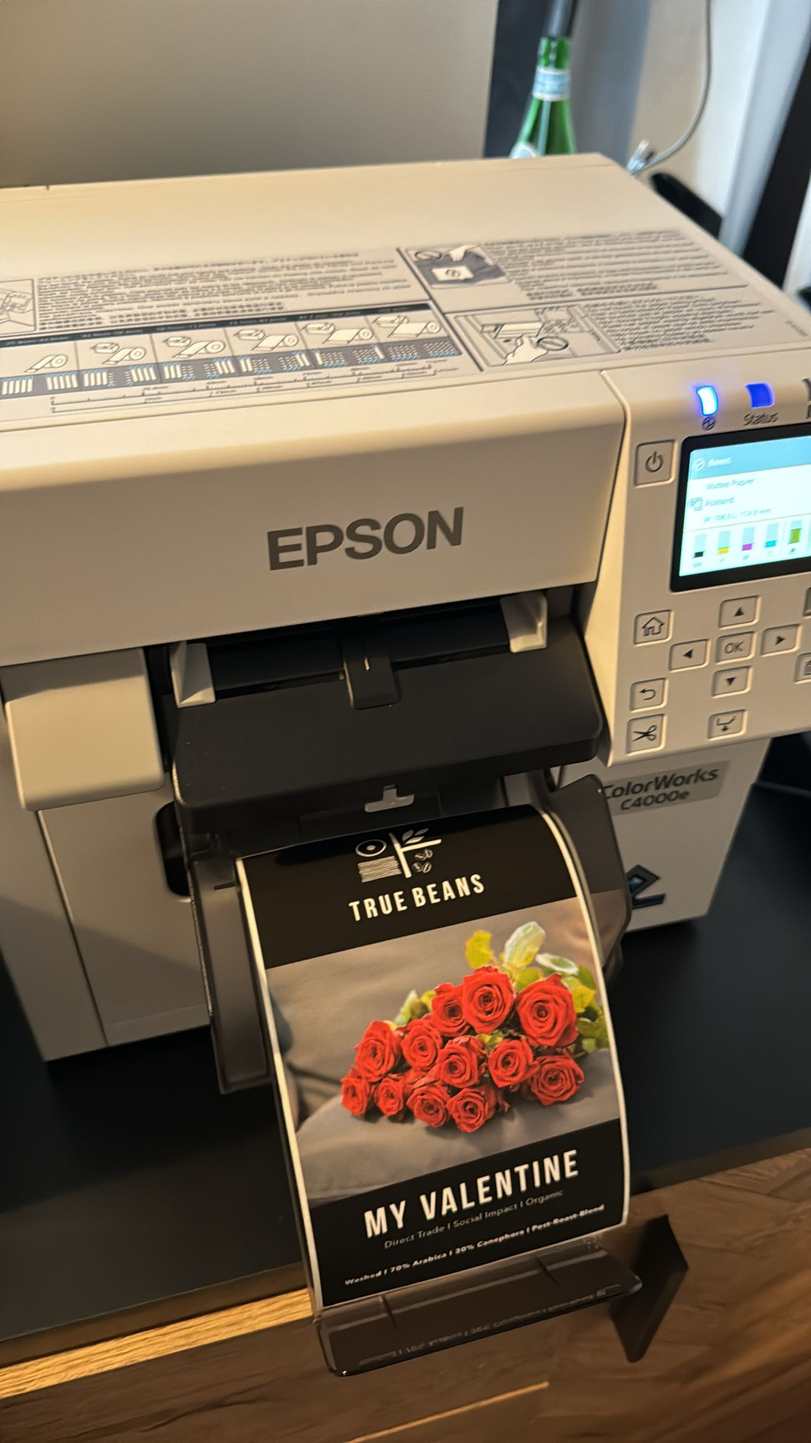 Ein Etikettendrucker von Epson druckt ein True Beans Kaffeeetikett mit der Aufschrift "My Valentine", das rote Rosen zeigt. Der Drucker steht auf einer Arbeitsfläche und das Etikett wird gerade aus dem Drucker ausgegeben. Im Hintergrund sind weitere Details des Druckers und eine grüne Flasche sichtbar.