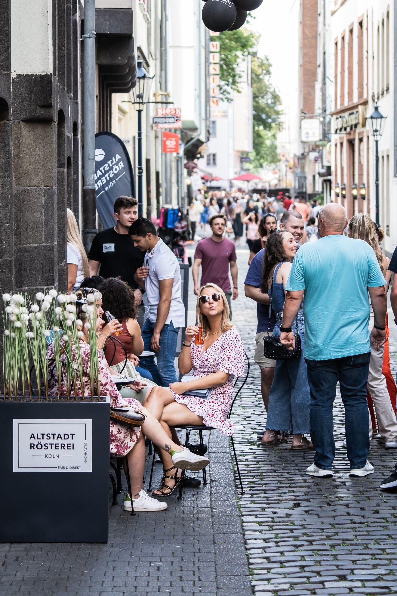 Eine belebte Straße in Köln, vor der Altstadt Rösterei sitzen Menschen an Tischen, trinken Kaffee und unterhalten sich. Im Hintergrund flanieren weitere Passanten.