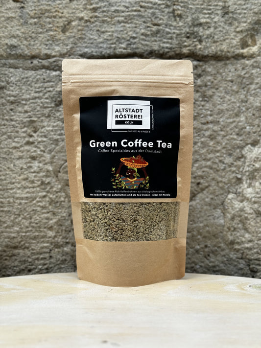 Eine Packung Altstadt Rösterei Green Coffee Tea vor einer Steinwand, mit der Aufschrift "100% granulierter Roh-Kaffeebohnen aus ökologischem Anbau" und einem Bild von Kaffeebohnen und Blättern.