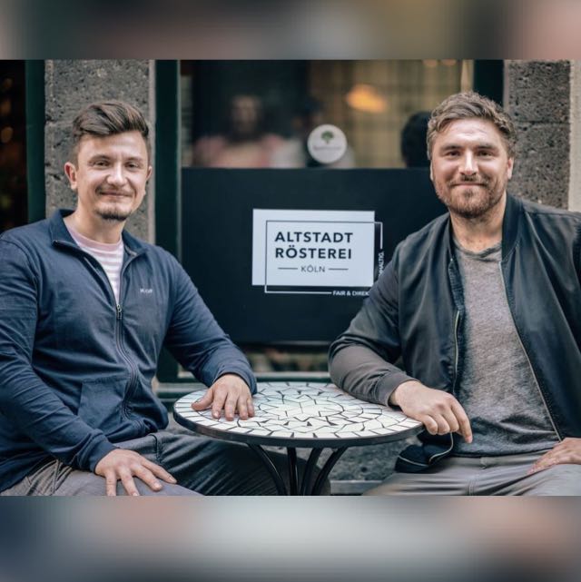 Die beiden Gründer Robin Kracht (links) und Patrick Köhler (rechts) sitzen vor ihrer Altstadt Rösterei in Köln an einem runden Tisch und lächeln in die Kamera.