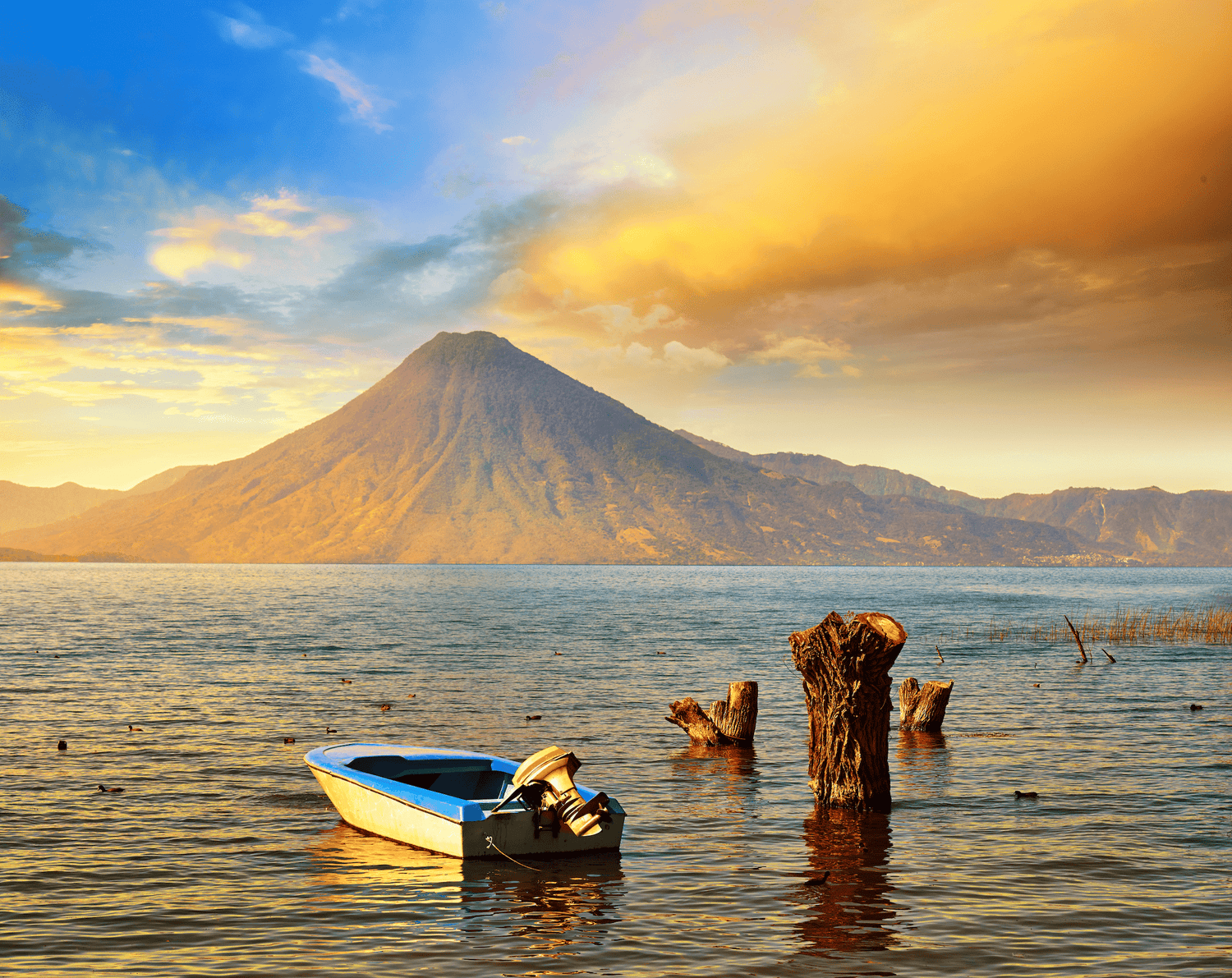 Ein malerischer Sonnenuntergang über dem Atitlán-See in Guatemala, mit einem ruhigen Boot im Vordergrund. Im Hintergrund erhebt sich majestätisch ein Vulkan, umgeben von Bergen, die in warmen, goldenen Farben des Sonnenuntergangs getaucht sind. Der Himmel ist eine Mischung aus Blau und Orange, was eine friedliche und idyllische Atmosphäre schafft.