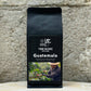 Ein Bild des Produkts Guatemala 100% Arabica von True Beans auf einem Holztisch vor einer steinernen Säule.