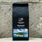 Ein Bild des Produkts Hawaii Kona Kaffee von True Beans auf einem Holztisch vor einer steinernen Säule.