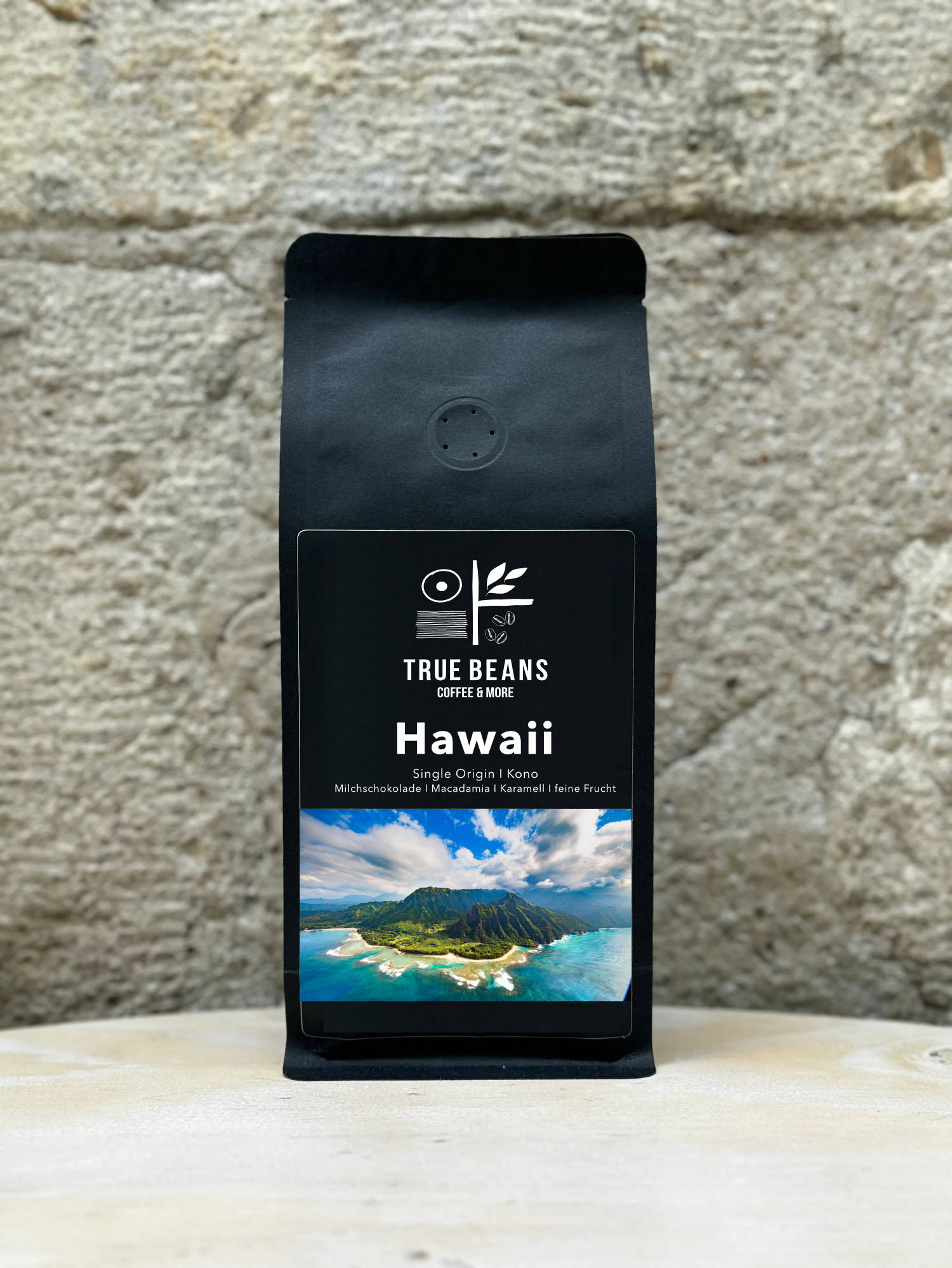Ein Bild des Produkts Hawaii Kona Kaffee von True Beans auf einem Holztisch vor einer steinernen Säule.