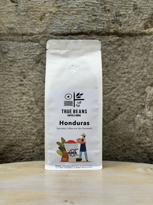 Eine weiße Packung True Beans "Honduras" Kaffee vor einer Steinwand. Auf dem Etikett ist ein stilisierter Kaffeebauer mit einem Wagen voller Kaffeekirschen abgebildet. Der Kaffee ist 100% Arabica, gewaschen verarbeitet, stammt aus der Kooperative Las Adelitas und wächst in einer Höhe von ca. 1.600 Metern. Der Kaffee wurde mit eigenem Solarstrom geröstet und hat eine SCA-Bewertung von 83,3.