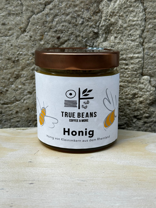 Ein Glas True Beans Honig vor einer Steinwand. Auf dem Etikett sind das True Beans Logo und Illustrationen von Bienen zu sehen, sowie die Beschreibung "Honig von Kleinimkern aus dem Rheinland".