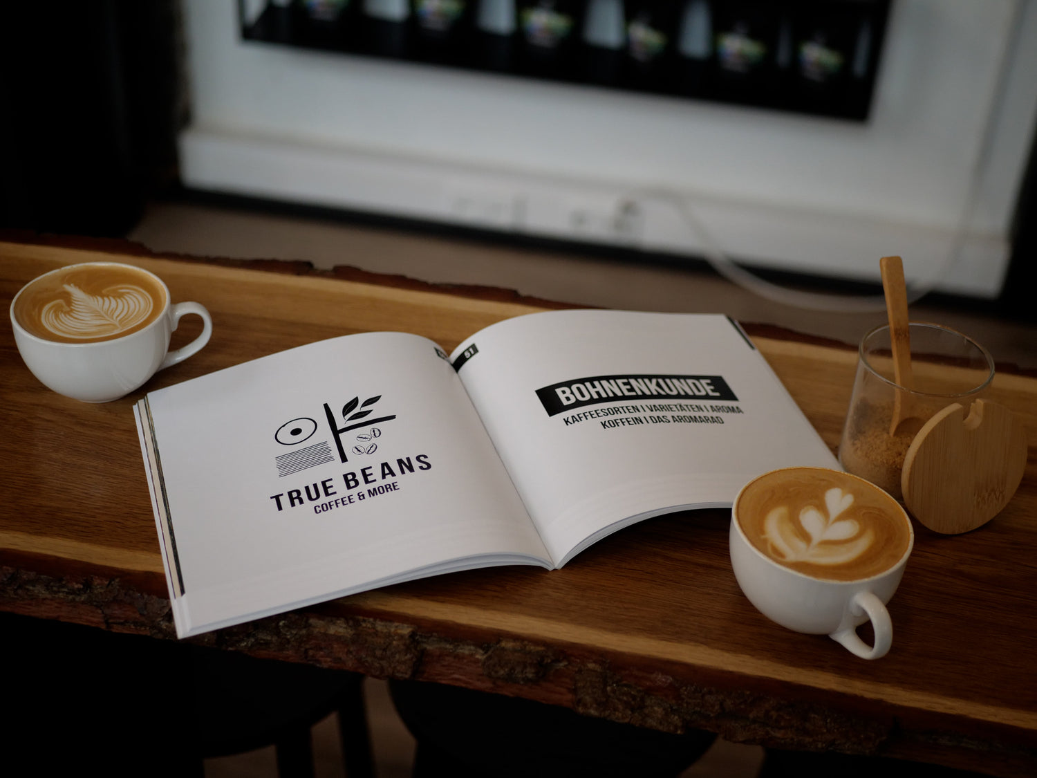 Ein aufgeschlagenes Buch mit dem Titel 'Bohnenkunde' liegt auf einem Holztresen, umgeben von zwei Tassen Kaffee mit Latte Art und einem Glas mit Holzdeckel, das Zucker enthält. Die Seite des Buches zeigt das Logo von 'True Beans Coffee & More'. Die Szene vermittelt eine gemütliche Atmosphäre in einem Café.