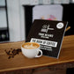 Ein Buch mit dem Titel 'The Book of Coffee' von 'True Beans Coffee & More' steht leicht geöffnet auf einem Holztresen, daneben eine Tasse Kaffee mit Latte Art und einige verstreute Kaffeebohnen. Die Szene vermittelt eine stilvolle, einladende Atmosphäre in einem Café.