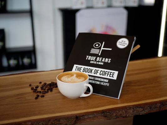 Ein Buch mit dem Titel 'The Book of Coffee' von 'True Beans Coffee & More' steht leicht geöffnet auf einem Holztresen, daneben eine Tasse Kaffee mit Latte Art und einige verstreute Kaffeebohnen. Die Szene vermittelt eine stilvolle, einladende Atmosphäre in einem Café.