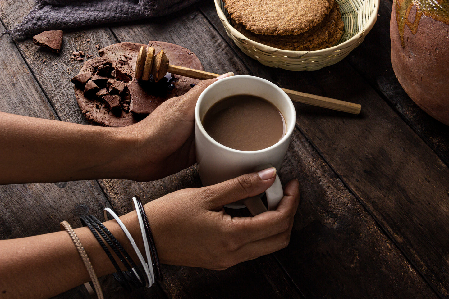 Ein Paar Hände hält eine weiße Tasse mit heißem Getränk, umgeben von einem rustikalen Holzbrett, auf dem sich eine Honigstange, zerbrochene Schokolade und ein Korb mit Keksen befinden. Die Szene ist gemütlich und lädt zum Genießen ein.