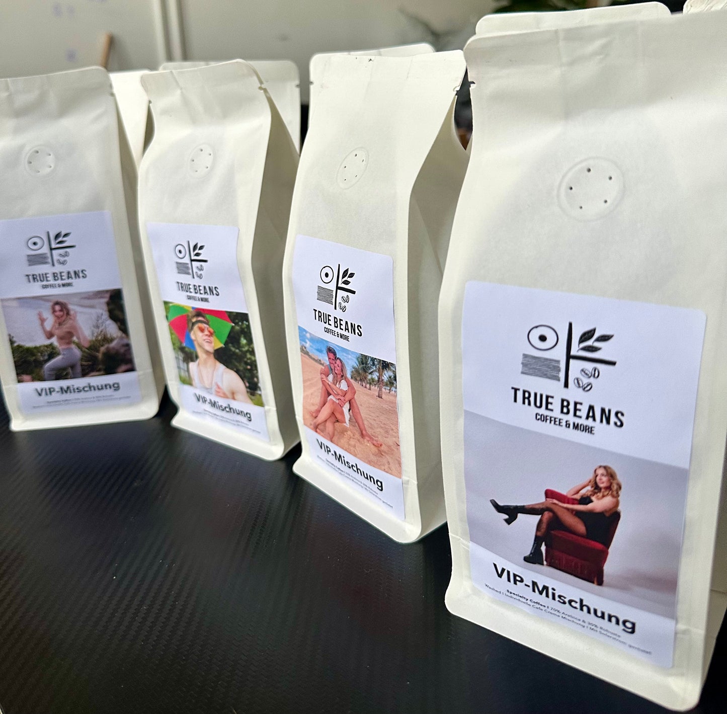 Mehrere Packungen True Beans VIP-Mischung Kaffee mit individuellen Etiketten, die verschiedene Personen in unterschiedlichen Umgebungen zeigen, stehen in einer Reihe auf einem Tisch.