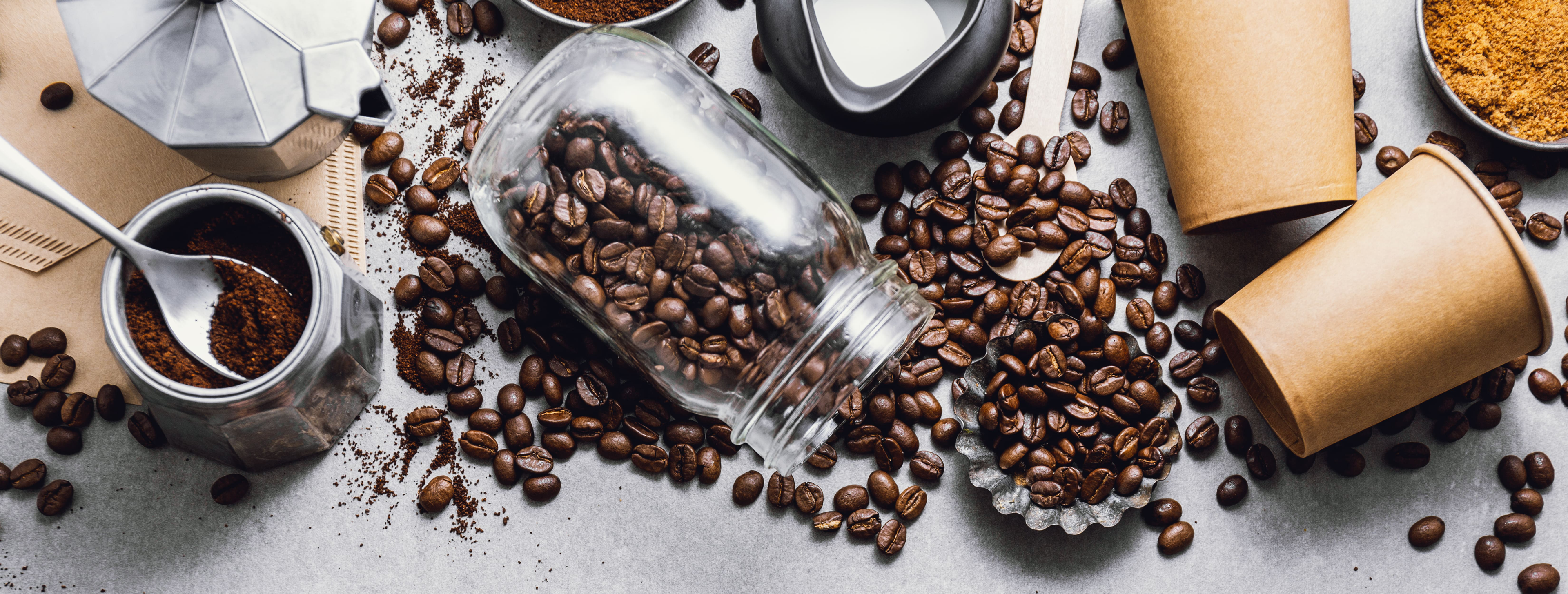 Verschiedene Kaffeebohnen, gemahlener Kaffee, eine Espressokanne und Pappbecher, arrangiert auf einer grauen Oberfläche, zeigen die Vielfalt der Kaffeezubereitung.