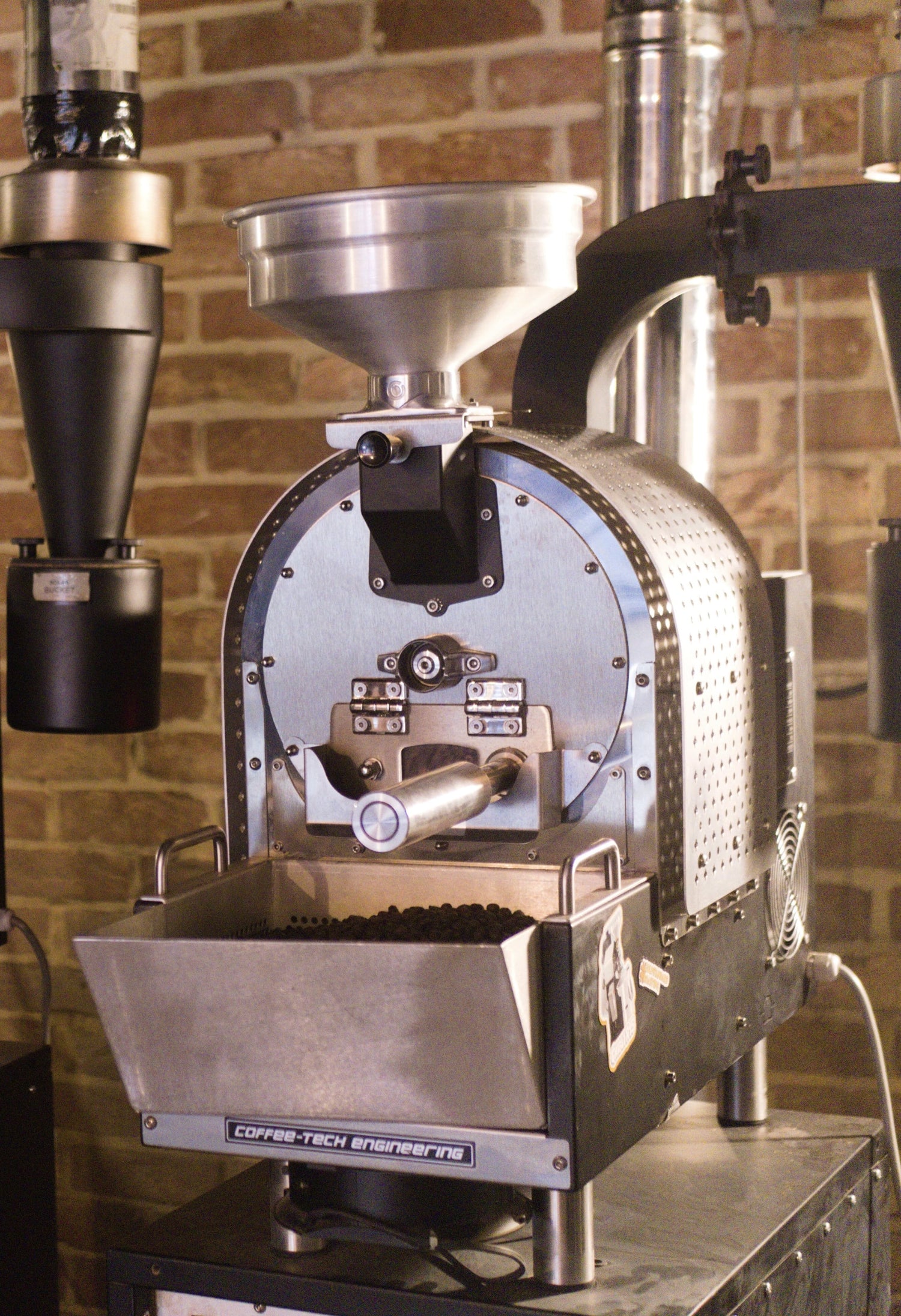 Eine Kaffeeröstmaschine aus Metall vor einer Backsteinwand, die gerade Kaffeebohnen röstet. Die Maschine hat einen Trichter oben und einen Behälter unten, in dem die gerösteten Bohnen gesammelt werden.