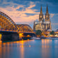 Nächtliche Ansicht der Hohenzollernbrücke und des Kölner Doms, beide beleuchtet, über dem ruhig fließenden Rhein unter einem wolkigen Abendhimmel.