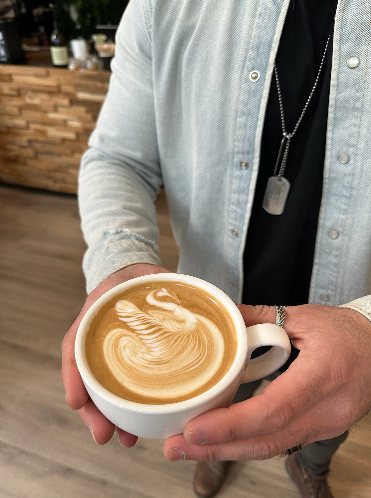 Eine Person hält eine Tasse Latte mit kunstvollem Schwan-Muster im Milchschaum, im Hintergrund ist ein Café zu sehen.