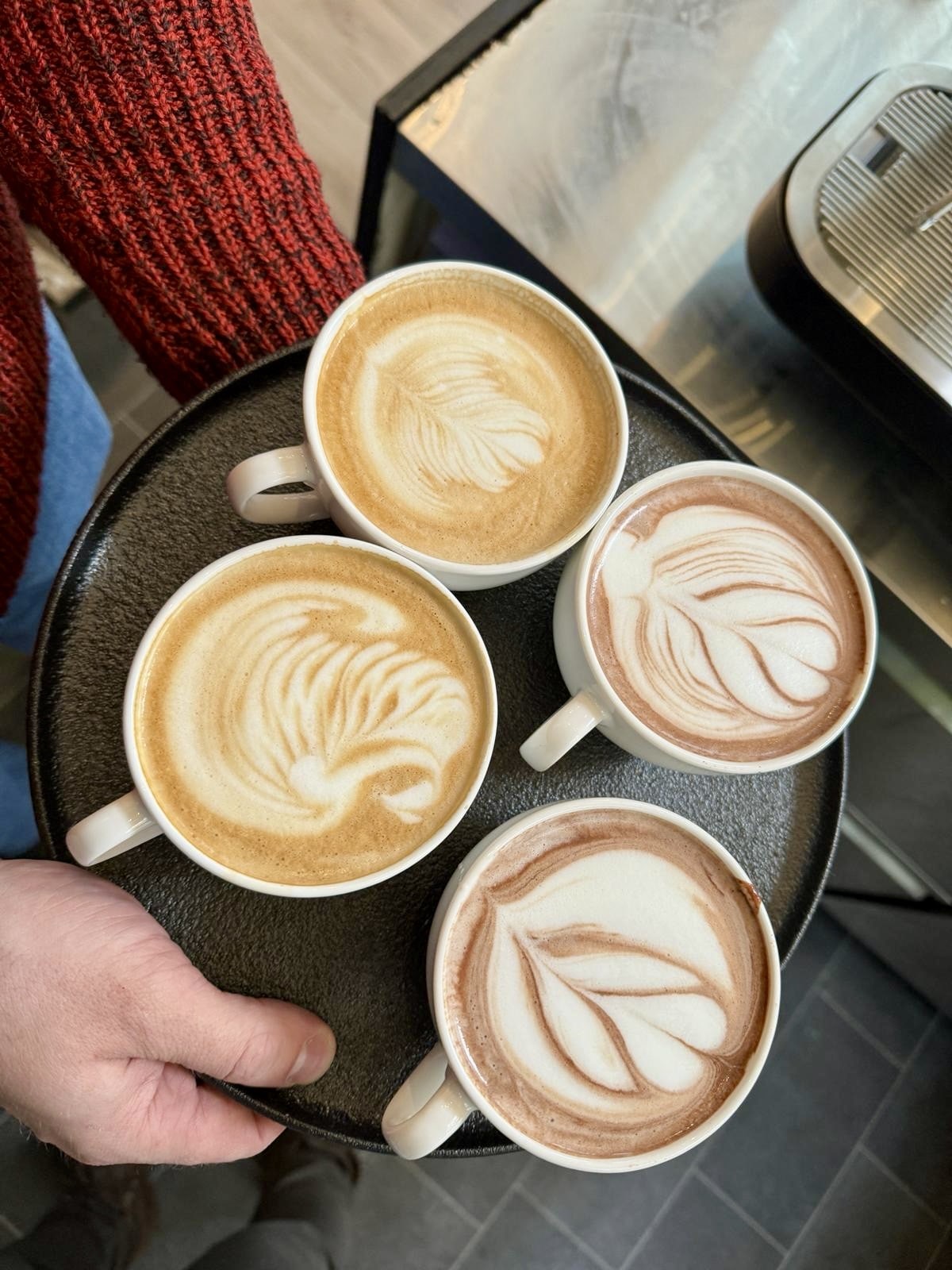 Eine Person hält ein Tablett mit vier Tassen Kaffee, die jeweils mit kunstvollen Mustern im Milchschaum verziert sind.