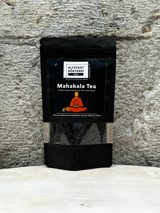 Eine Packung Altstadt Rösterei Köln Mahakala Tea vor einer Steinwand. Auf der Verpackung ist eine Illustration eines meditierenden Mönchs sowie die Beschreibung "Coffee Specialties aus der Domstadt" zu sehen.