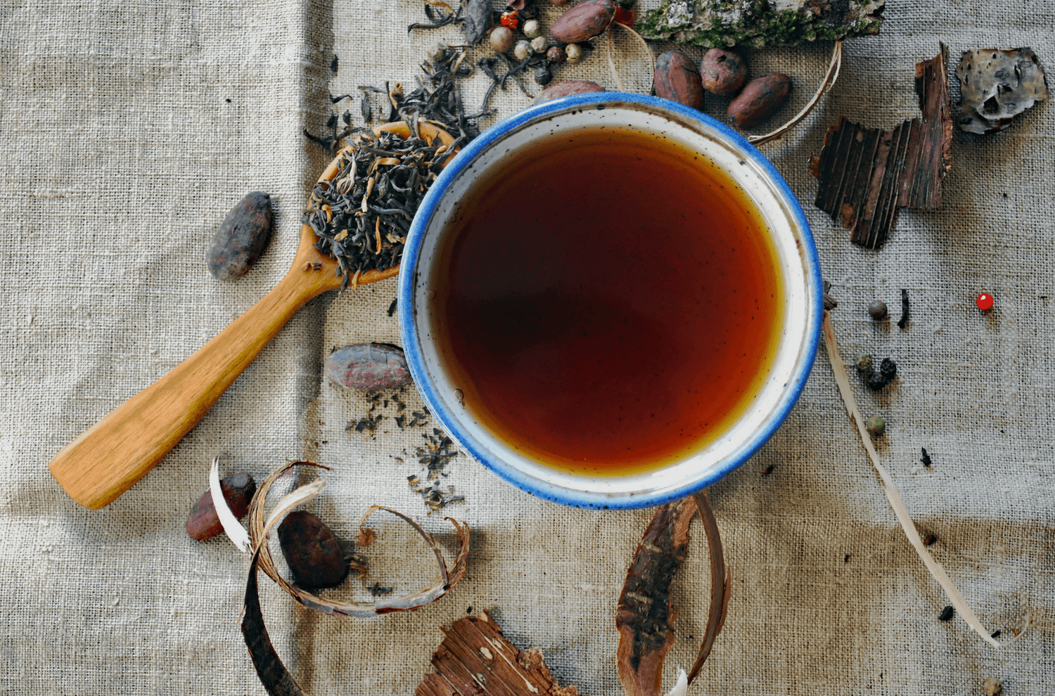 Eine Tasse mit dunkelbraunem Tee steht auf einer Leinendecke, umgeben von verschiedenen Teeblättern, Kakaobohnen, Holzstücken und Gewürzen. Ein Holzlöffel mit losen Teeblättern liegt daneben, was eine natürliche und rustikale Atmosphäre vermittelt.