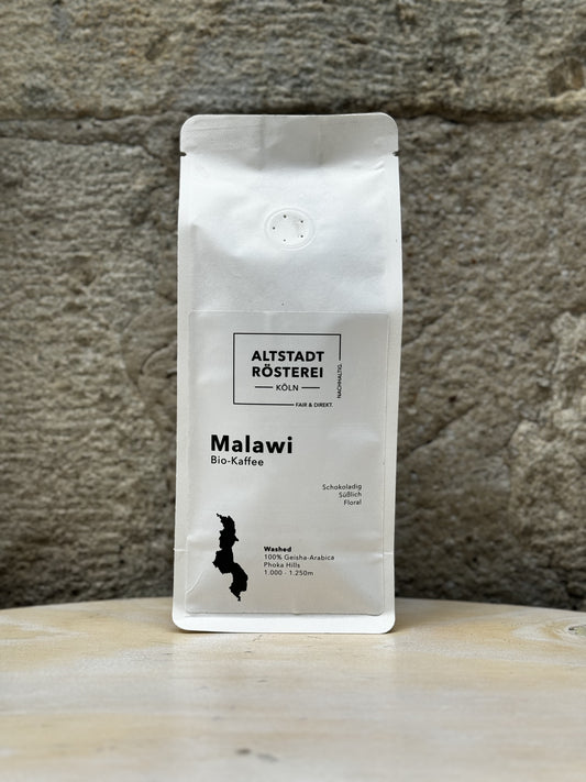Eine weiße Packung Altstadt Rösterei "Malawi Bio-Kaffee" vor einer Steinwand, mit der Aufschrift "Fair & Direkt". Auf dem Etikett ist eine stilisierte Karte von Malawi abgebildet. Der Kaffee ist 100% Geisha-Arabica aus den Phoka Hills, gewaschen verarbeitet und wächst in Höhen von 1.000 bis 1.250 Metern. Die Geschmacksbeschreibung lautet: schokoladig, süßlich, floral.