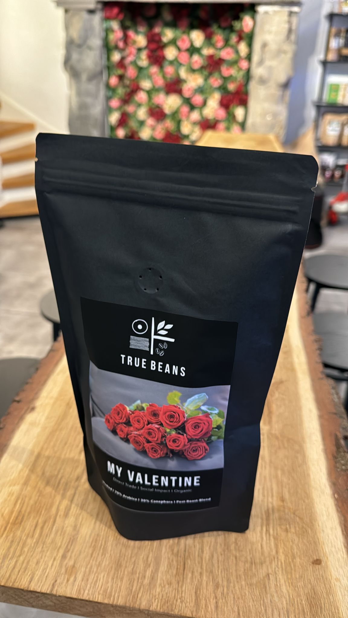 Eine Packung True Beans Kaffee "My Valentine" steht auf einem Holztisch. Die Verpackung zeigt ein Bild von roten Rosen und der Text auf der Packung beschreibt den Kaffee als Direct Trade, Social Impact und Organic. Im Hintergrund sind unscharf Blumentapeten und Regale zu sehen.