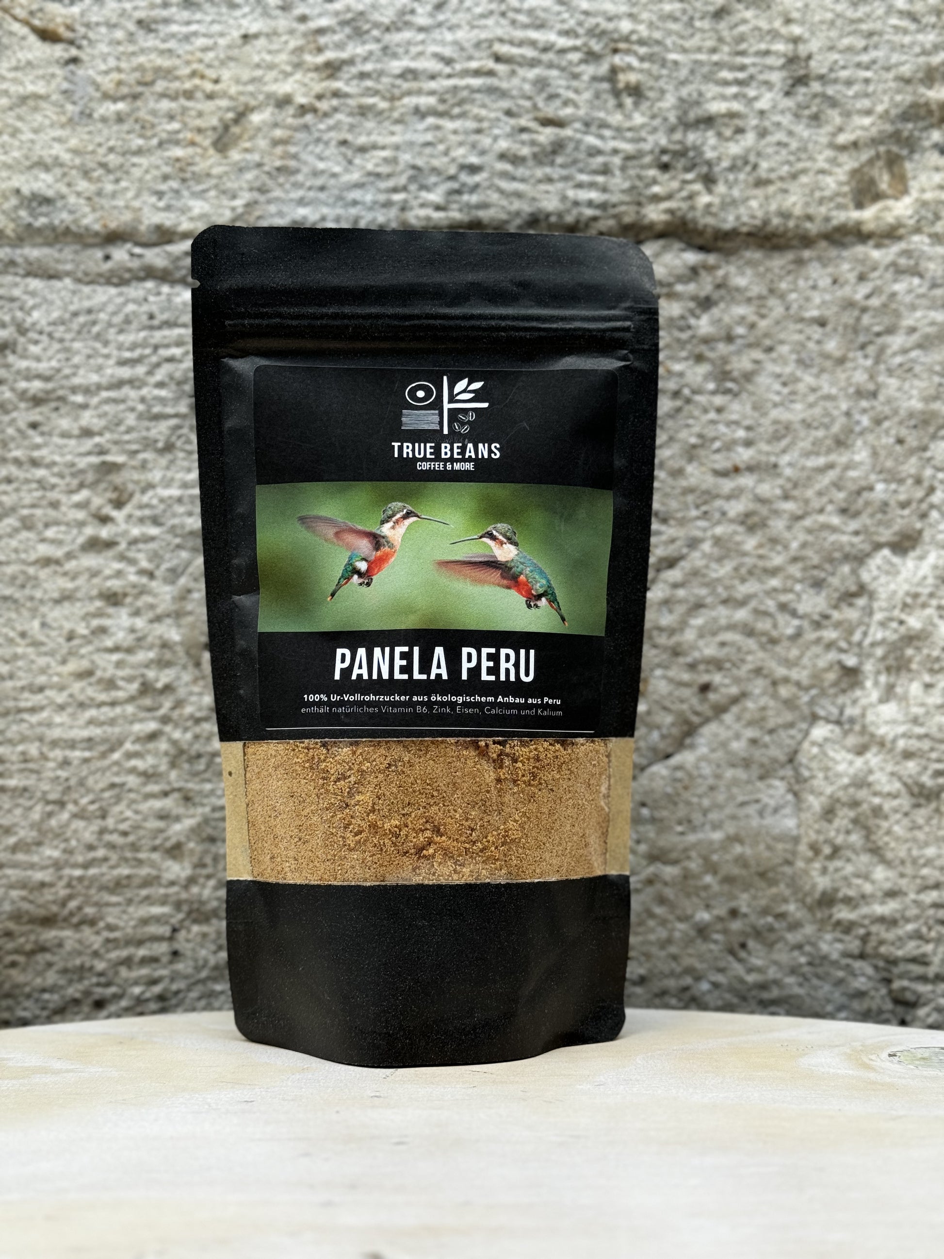 Eine Packung True Beans Panela Peru vor einer Steinwand, mit der Aufschrift "100% Ur-Vollrohrzucker aus ökologischem Anbau aus Peru" und einem Bild von zwei Kolibris, die sich gegenüberstehen.