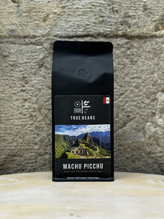 Eine Packung True Beans Kaffee "Machu Picchu" vor einer Steinwand, mit der Aufschrift "Direct Trade | Andenkaffee | Organic | Peru". Auf dem Etikett ist eine malerische Ansicht von Machu Picchu mit den Anden im Hintergrund abgebildet. Der Kaffee ist 100% Arabica, gewaschen verarbeitet und stammt aus einem einzigen Ursprungsgebiet.